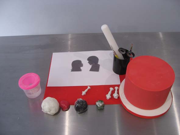 Matériel : 1 gâteau de 15 cm de diamètre recouvert de pâte à sucre rouge. 2 patrons silhouettes «Homme» et «Femme». Découpoirs cœurs de tailles différentes. Gumpaste noire, blanche, rouge, verte.  1 petit rouleau 1 cutter 1 pinceau Eau